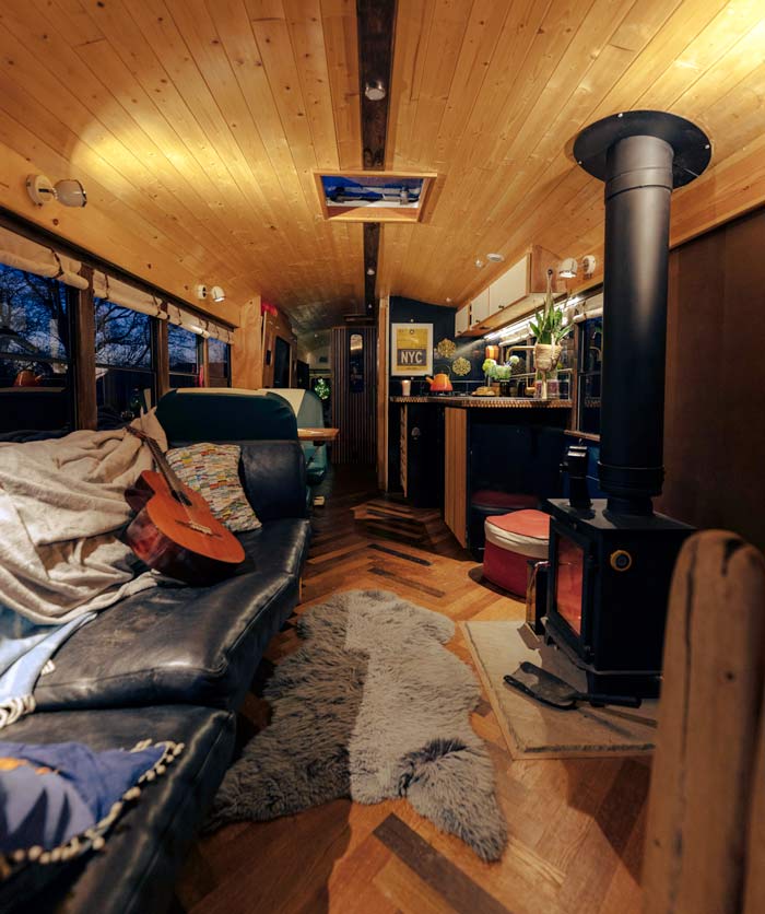 Warm and inviting interior of the Skoolie Stays glamping bus with wood-panelled ceiling, log-burning stove, leather sofa, guitar, and stylish kitchen area with ambient lighting.