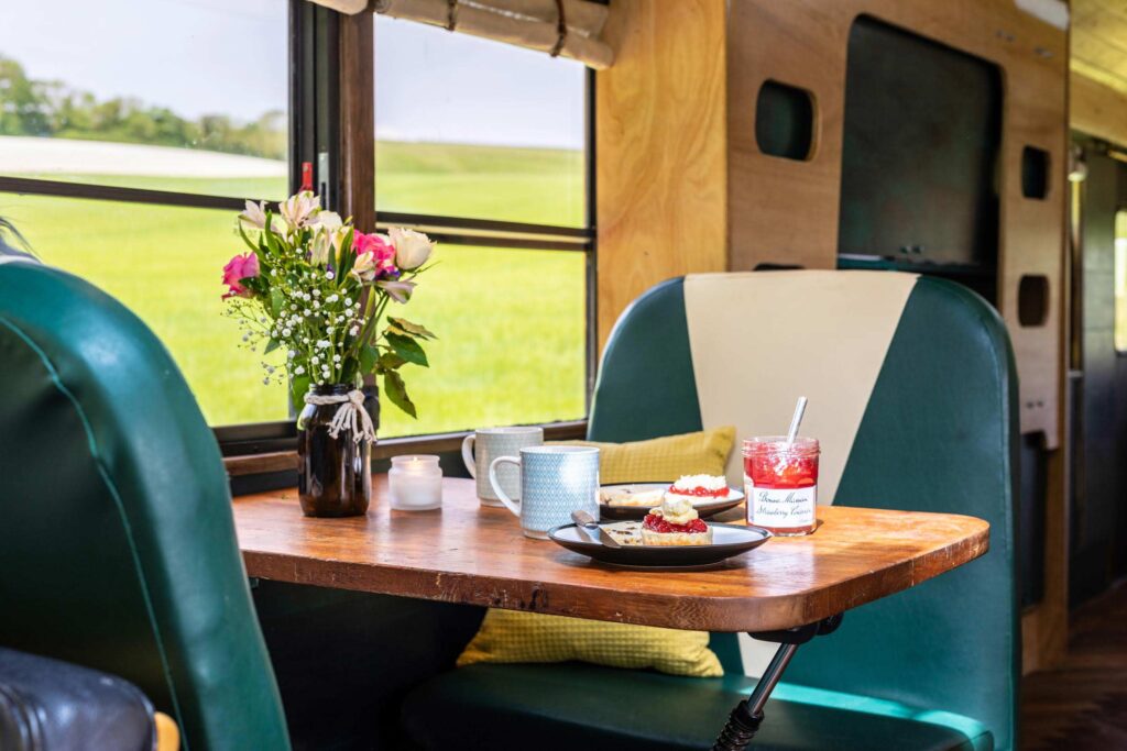 Vintage diner-style seating inside the Skoolie Stays glamping bus with fresh flowers, cream tea, mugs, and strawberry conserve on a wooden table overlooking green Sussex fields.
