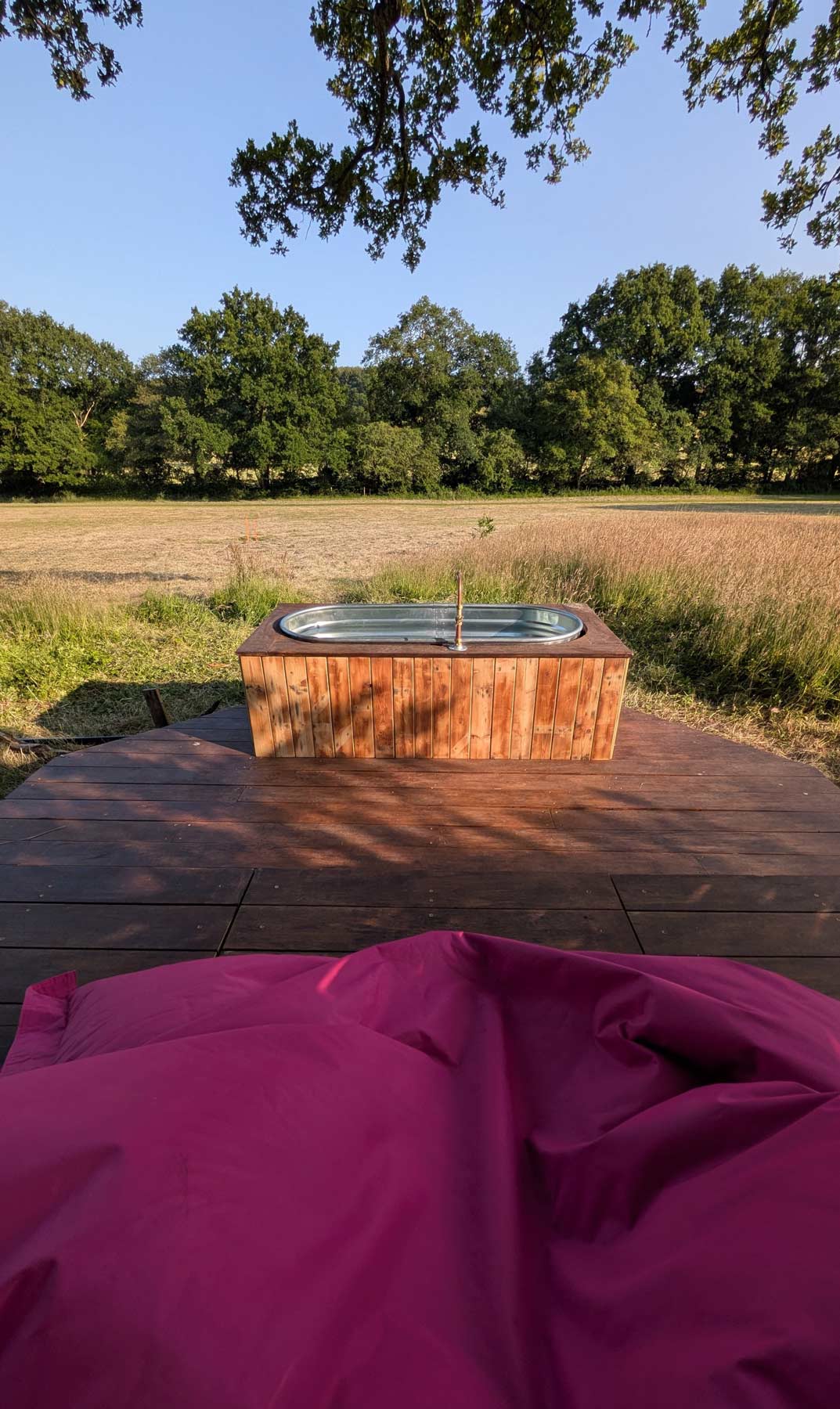 View from a pink bean bag on the deck at Skoolie Stays, looking out to a wood-fired hot tub and peaceful meadow surrounded by trees under a clear blue sky.