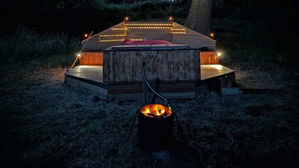 Cozy nighttime glamping scene with fairy lights, wooden deck, and hot tub beneath large oak tree at Skoolie Stays.
