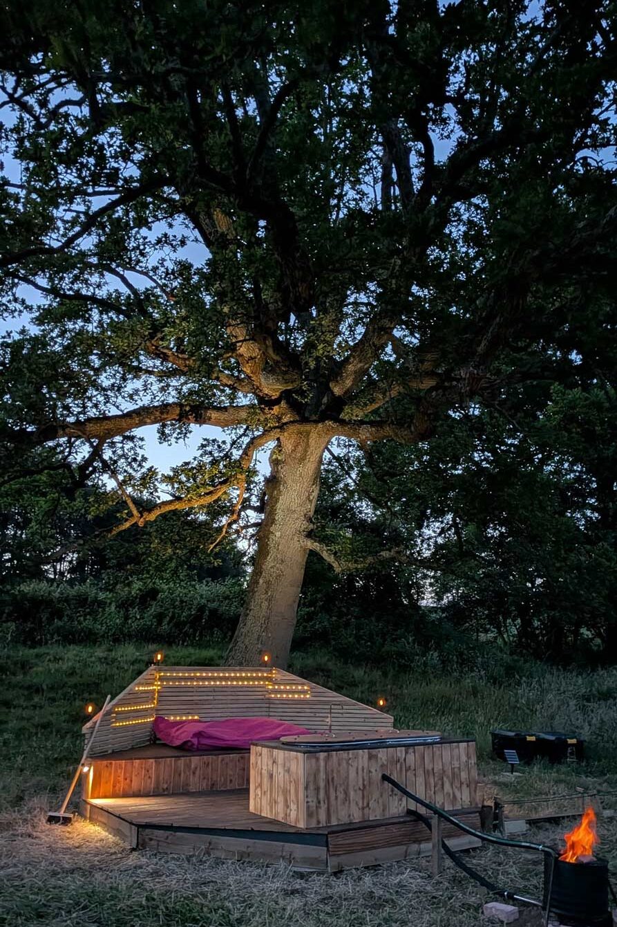 Cozy nighttime glamping scene with fairy lights, wooden deck, and hot tub beneath large oak tree at Skoolie Stays converted glamping school bus