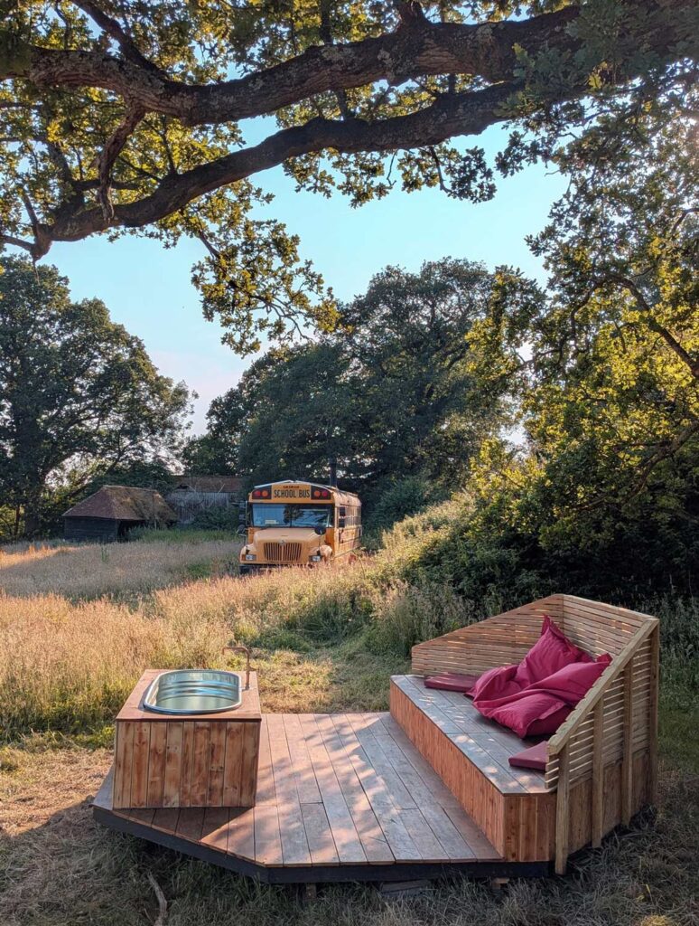 Wood-fired hot tub and cosy deck with outdoor seating and pink cushions under a large oak tree, with Skoolie Stays yellow glamping bus in the background surrounded by wild grass and trees in the Sussex countryside