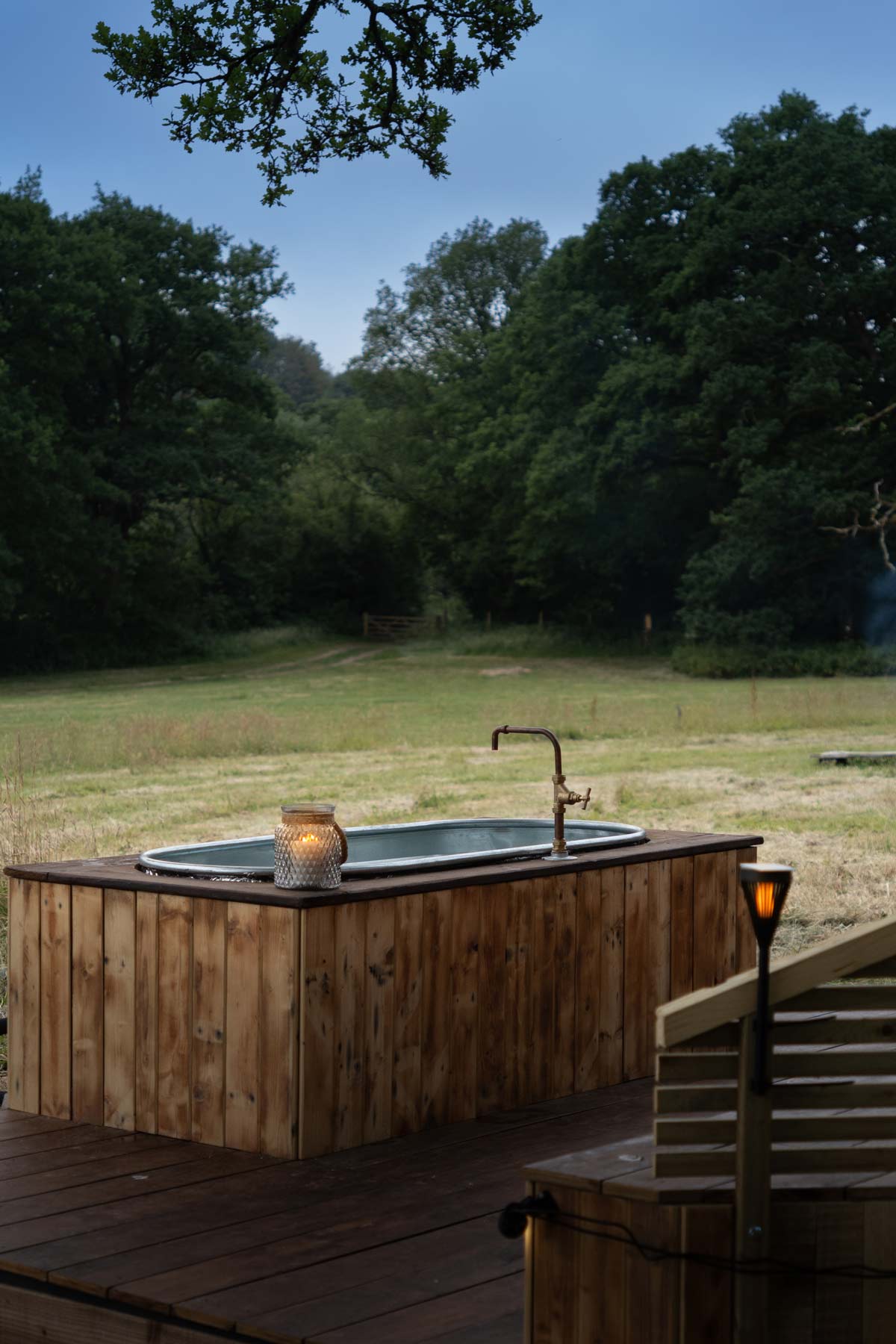 Wood-fired hot tub glowing by candlelight in peaceful countryside field at dusk, surrounded by trees at Skoolie Stays american school bus