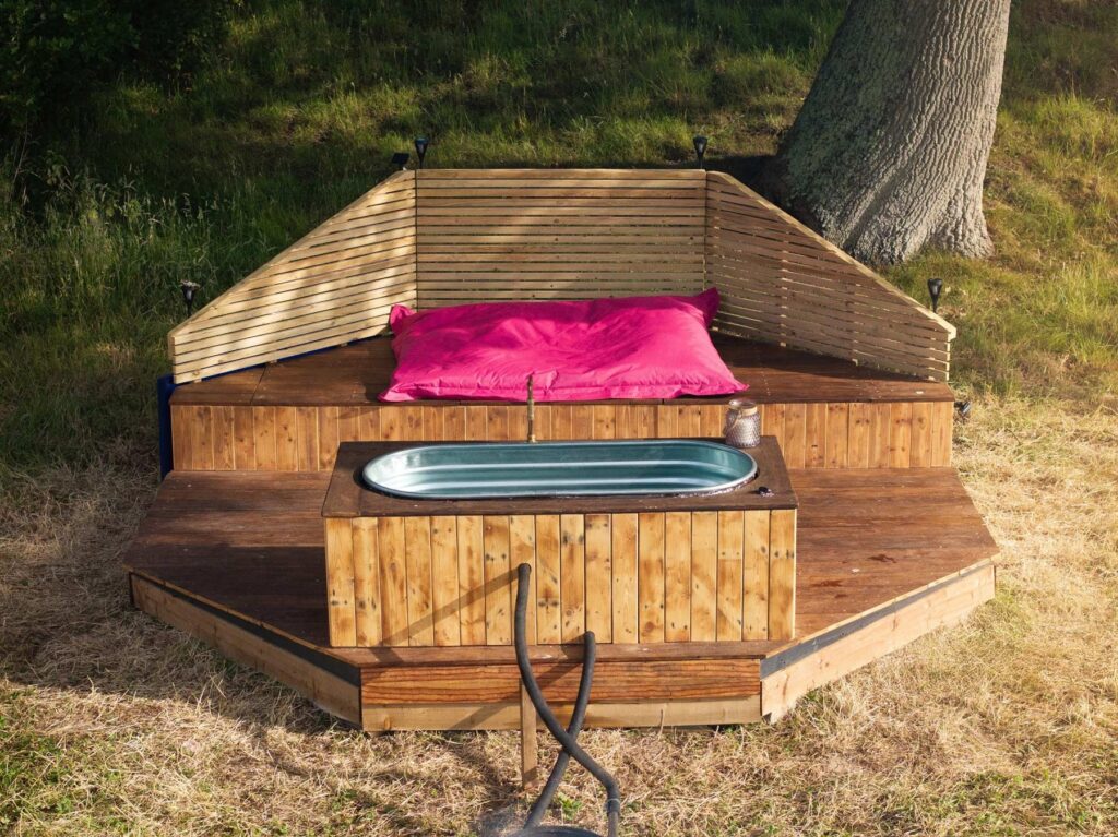 Private glamping hot tub setup with bench and cushions nestled under mature oak tree at Skoolie Stays retreat.