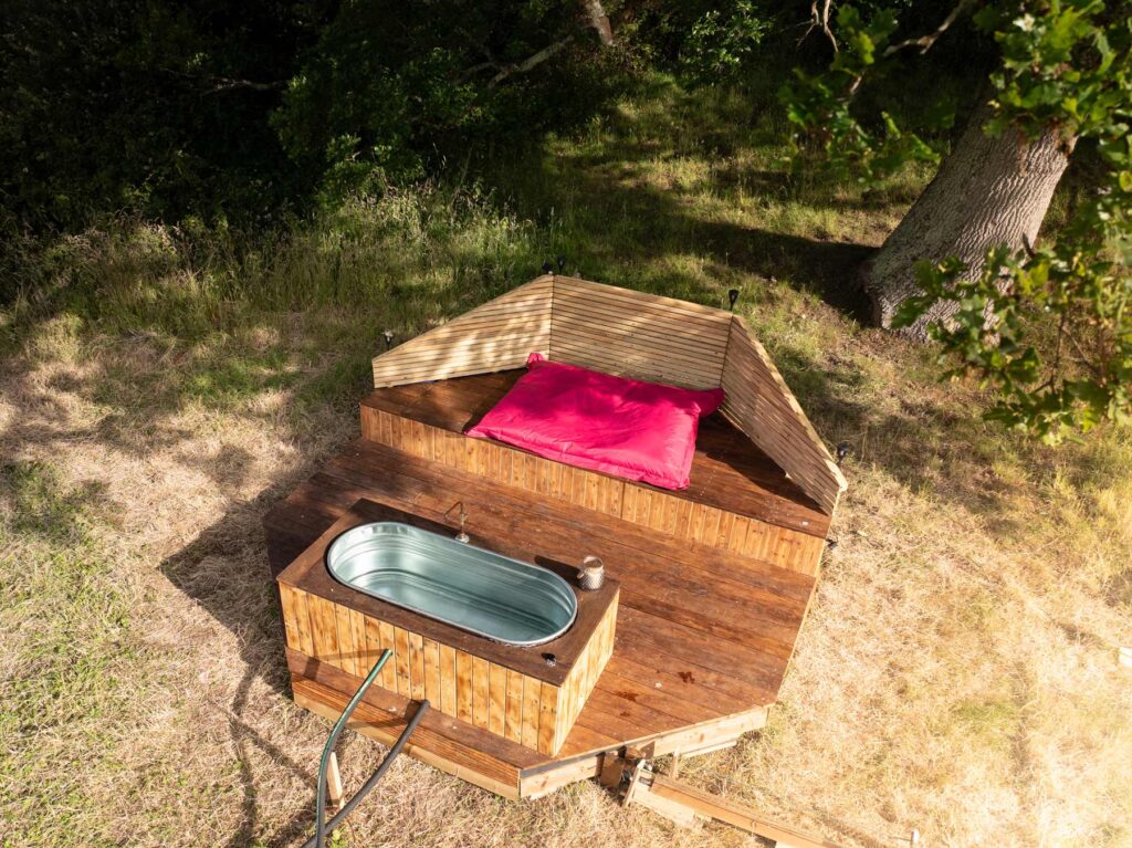 Aerial view of hot tub setup with seating area and large pink bean bag under oak tree at Skoolie Stays glamping site.
