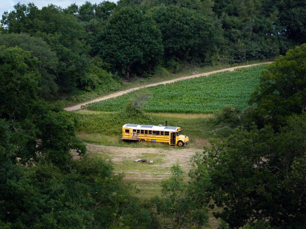 A converted yellow American school bus set up as a glamping accommodation, parked in a secluded green field surrounded by trees and countryside