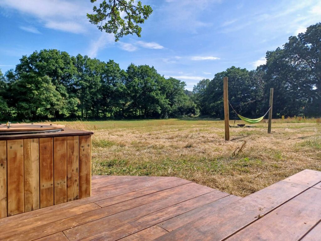 View from wooden deck with hot tub at Skoolie Stays glamping site, looking across a peaceful Sussex meadow with a hammock strung between posts and surrounded by trees under a blue summer sky.