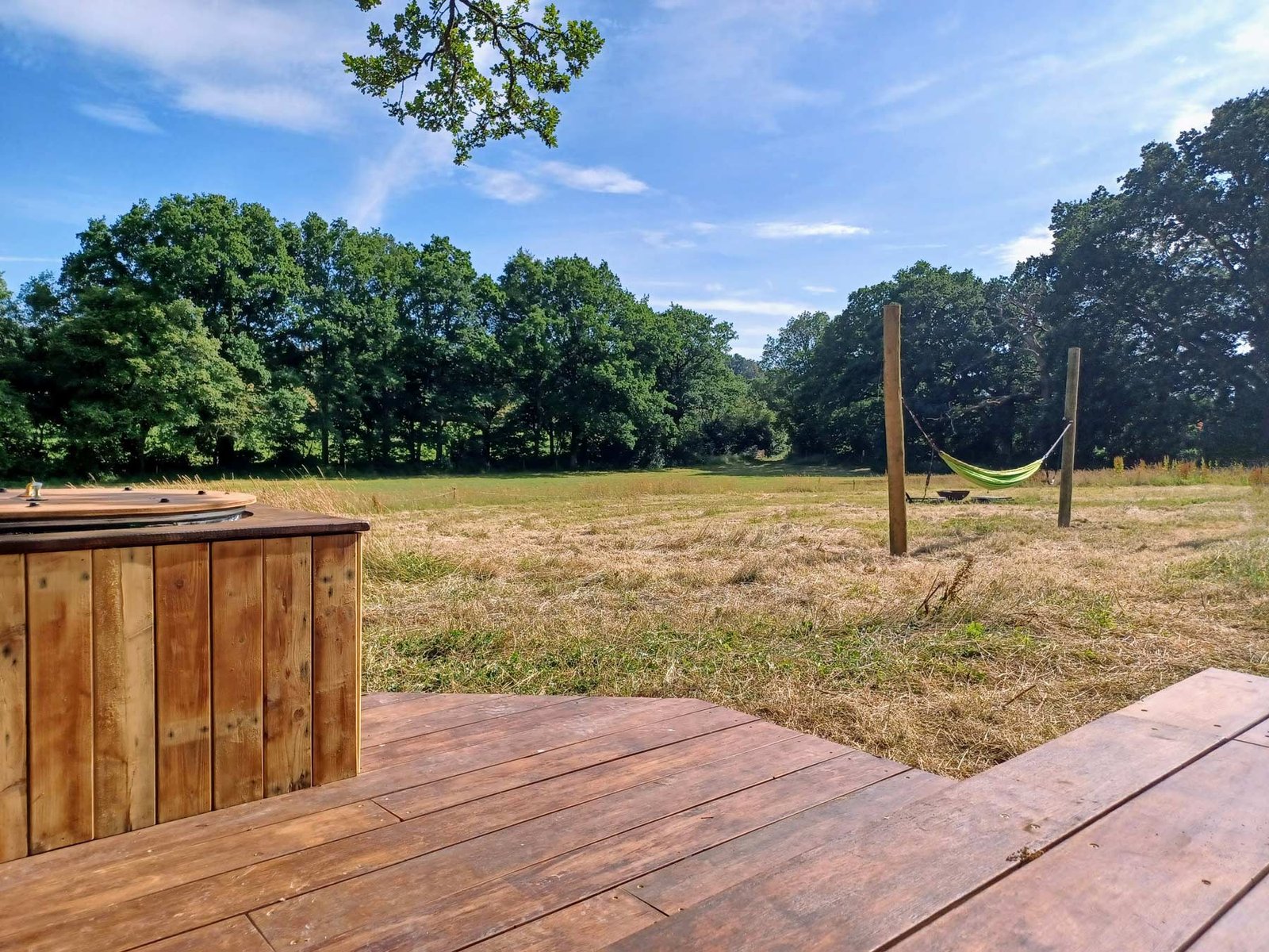 View from wooden deck with hot tub at Skoolie Stays glamping site, looking across a peaceful Sussex meadow with a hammock strung between posts and surrounded by trees under a blue summer sky.