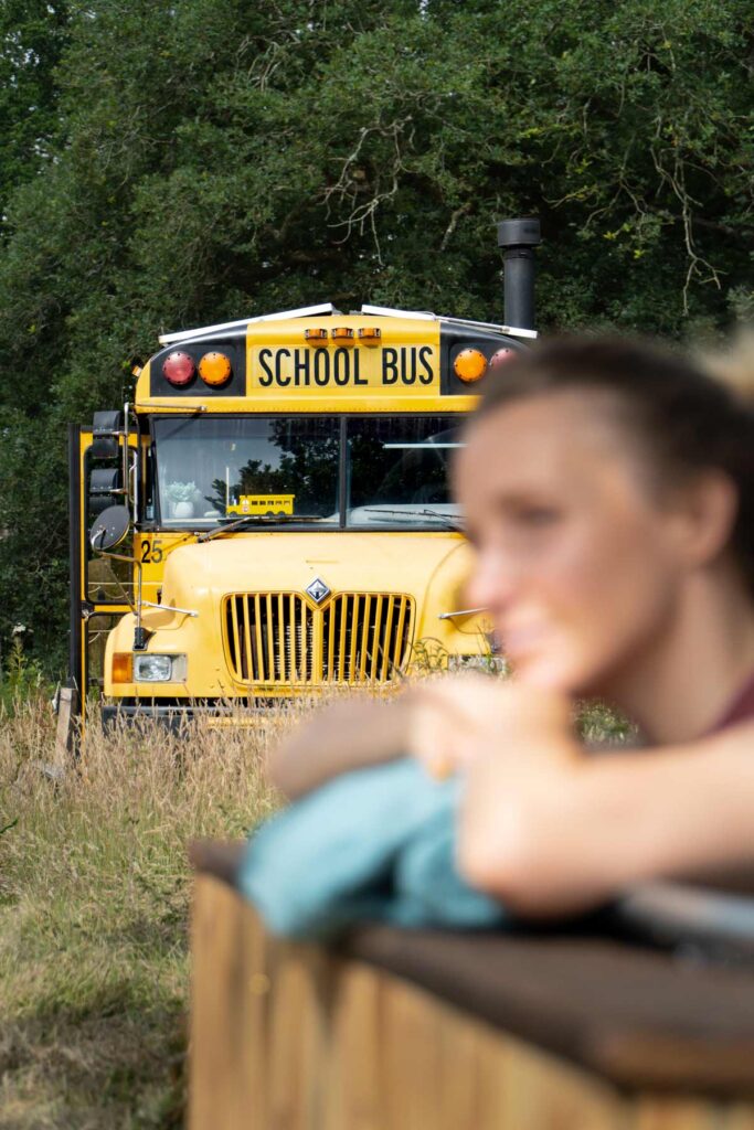 Woman relaxing in a wood-fired hot tub with a vintage American school bus in the background at Skoolie Stays glamping site in West Sussex.
